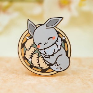May include: A gray enamel pin featuring a cartoon character with a bunny-like appearance, resting on a bed of dumplings. The pin has a circular design with a gold border and star accents. The character has a sweet expression.