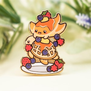 May include: A gold-colored enamel pin featuring a cartoon turtle with a pancake shell and a stack of pancakes on its head. The turtle is decorated with blueberries and strawberries, and it stands on a white plate.