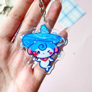 Op de afbeelding: Een heldere acryl sleutelhanger met een stripfiguur met een blauwe hoed, rode sjaal en een ster-vormige toverstaf. De sleutelhanger is bevestigd aan een zilveren sleutelhanger en ketting. Het karakter heeft een knipogende uitdrukking.