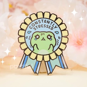 May include: A pin featuring a cartoon frog with a stressed expression inside a blue bubble, surrounded by a yellow rosette with the words "CONSTANTLY STRESSED". The pin has blue and yellow ribbons.