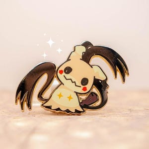May include: A Mimikyu enamel pin featuring a cream-colored ghost with black accents and a cheerful expression. The pin has a gold outline and two yellow stars on its body. The design is a stylized representation of a popular character.