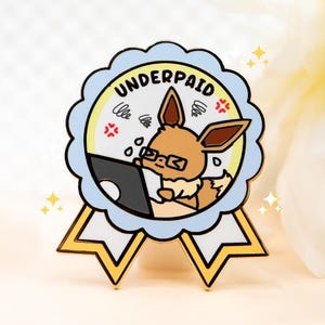 May include: A circular enamel pin shaped like a ribbon award with a light blue scalloped border. The centre features a brown cartoon creature with glasses working on a laptop, with the text UNDERPAID arched above and small yellow star accents.