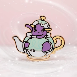 Puede incluir: Un pin de esmalte caprichoso que presenta una criatura púrpura dentro de una tetera blanca y dorada. La criatura tiene una cara sonriente y viste un atuendo verde claro. El pin tiene detalles dorados y está sobre un fondo rosa suave.