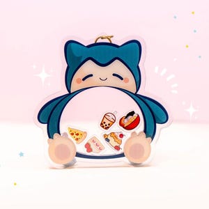 Peut inclure: Un porte-clés shaker en acrylique bleu et crème en forme de créature endormie, doté d'un ventre transparent contenant de petites breloques mobiles représentant divers aliments comme une pizza, un thé boba, un bol de ramen et un gâteau.