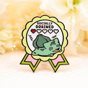 May include: A circular enamel pin shaped like a ribbon award with a light green border and pink accents. The center features a light green creature with a bulb on its back, the text "SOCIALLY DRAINED," and a row of five pixelated heart icons.