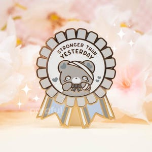 May include: A circular enamel pin shaped like a ribbon rosette with a light blue and grey border. The centre features a grey bear with a bandage on its head and the text STRONGER THAN YESTERDAY. Two ribbon tails hang from the bottom of the pin.