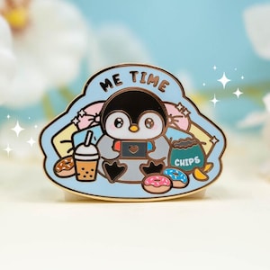 May include: A colorful enamel pin featuring a cartoon penguin playing a handheld video game. The pin has the words "ME TIME" and includes images of snacks like donuts, chips, and a drink. The penguin is black, white, and gray.