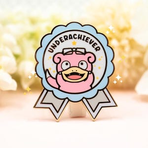 May include: A circular enamel pin shaped like a ribbon award with a light blue scalloped border and grey tails. The center features a pink cartoon creature wearing glasses with the text UNDERACHIEVER arched above in black capital letters.