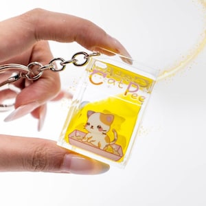 Può includere: Un portachiavi rettangolare in acrilico trasparente riempito con liquido giallo e glitter. La parte anteriore presenta un'illustrazione a cartone animato di un gatto bianco e arancione in una lettiera con la scritta Cat Pee in lettere viola.