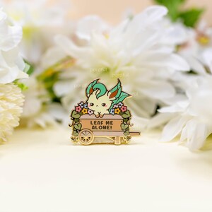 May include: A gold enamel pin depicting a cartoon character with green and cream features, in a cart decorated with flowers. The cart has a sign that reads "LEAF ME ALONE!"