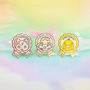 May include: Three enamel pins with gold trim and ribbon details. The pins feature cartoon characters and text: "ONE TOUGH PUFF", "CRYBABY", and "OVERTHINKER". The pins are in shades of pink, yellow, and cream, with a pastel background.