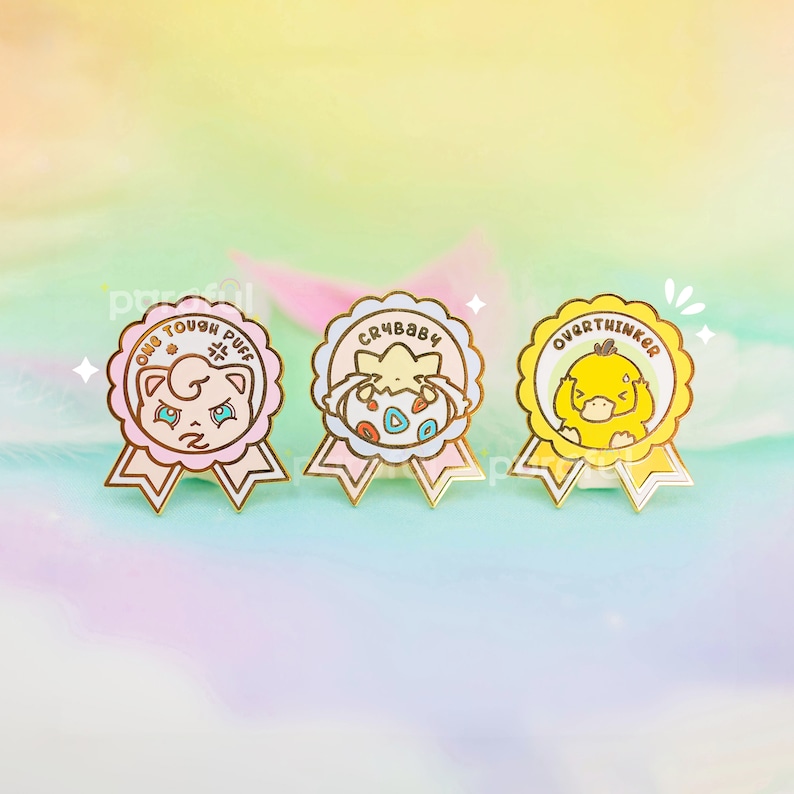 May include: Three enamel pins with gold trim and ribbon details. The pins feature cartoon characters and text: "ONE TOUGH PUFF", "CRYBABY", and "OVERTHINKER". The pins are in shades of pink, yellow, and cream, with a pastel background.