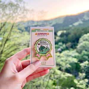 May include: A circular enamel pin shaped like a ribbon badge featuring a green creature with a bulb on its back, the text "SOCIALLY DRAINED," and a heart icon. The pin is mounted on a white backing card with the text "ERROR" and "SOCIAL BATTERY: 0%."