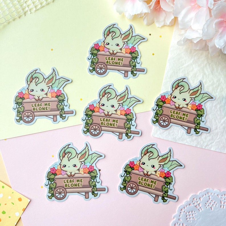 May include: A collection of six stickers featuring a cartoon creature with green leaf-like ears and wings, nestled in a wooden cart filled with flowers. The cart has the text "LEAF ME ALONE!". The stickers are on a pastel background.