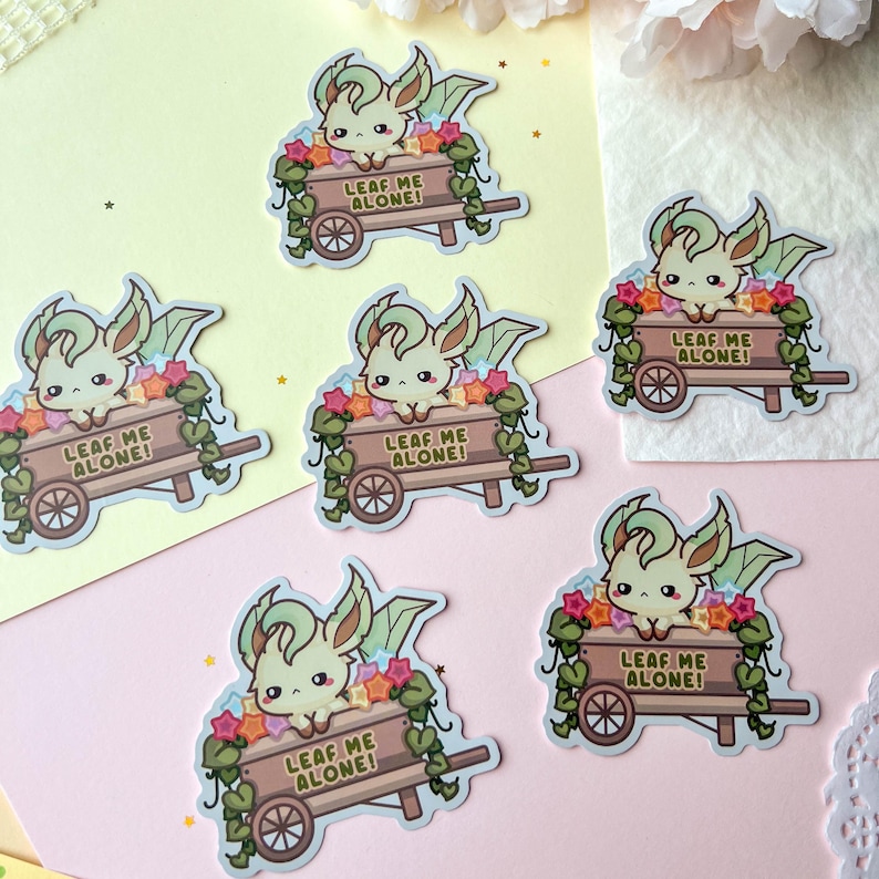 May include: A collection of stickers featuring a cartoon creature with green and cream-colored fur, large ears, and a small face. The creature is in a wooden cart filled with colorful stars and vines. The text "LEAF ME ALONE!" is on the cart.