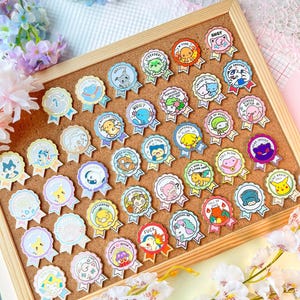 May include: A collection of colorful enamel pins displayed on a corkboard. Each pin features a cartoon character or design within a ribbon-shaped frame, with various phrases like "STRESSED" and "FUCK". The pins are set against a backdrop of flowers.