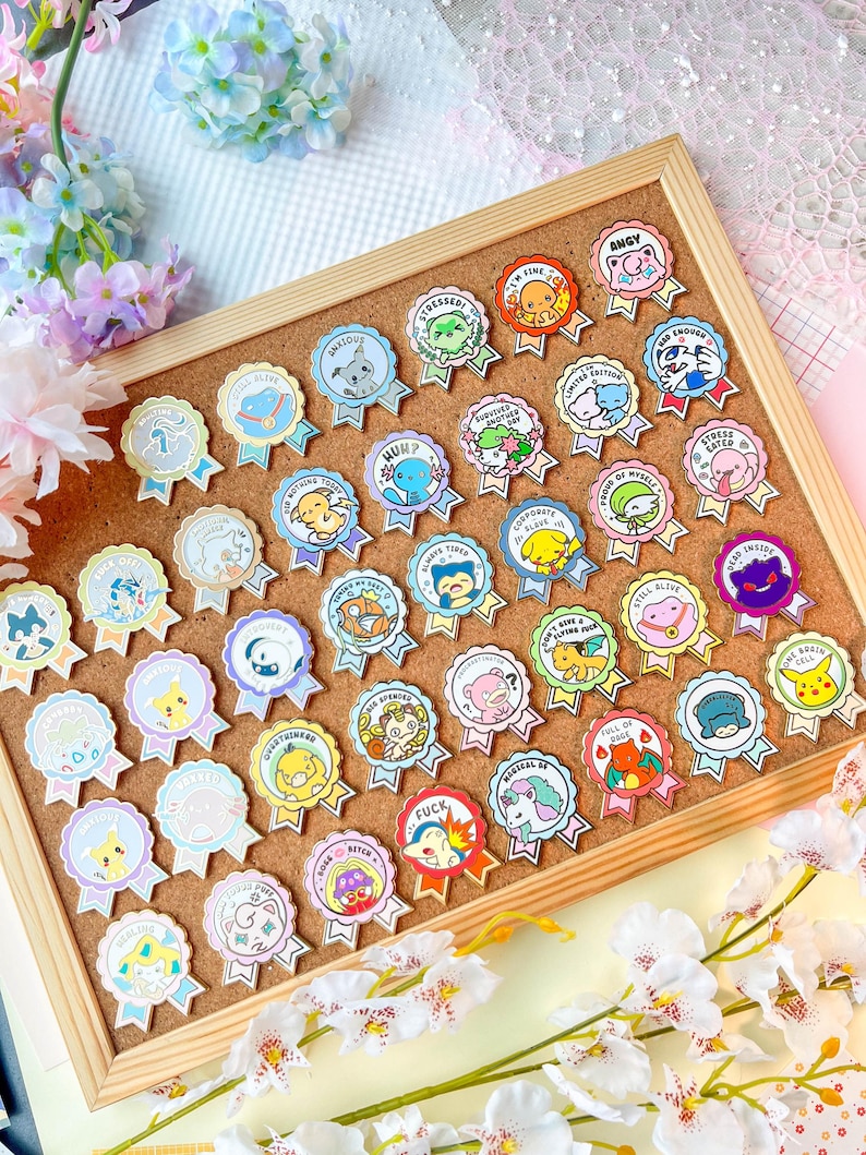 May include: A collection of colorful enamel pins displayed on a corkboard. Each pin features a cartoon character or design within a ribbon-shaped frame, with various phrases like "STRESSED" and "FUCK". The pins are set against a backdrop of flowers.