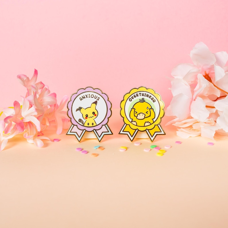 May include: Two enamel pins with cartoon characters. One pin is lavender with a yellow character and the word "ANXIOUS". The other is yellow with a yellow character and the word "OVERTHINKER". Both pins have a ribbon design.