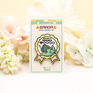 May include: A metal enamel pin shaped like a scalloped award ribbon with pink and light green accents. It features a green creature illustration, the text "SOCIALLY DRAINED," and a pixelated heart icon. The pin is mounted on a white display card.