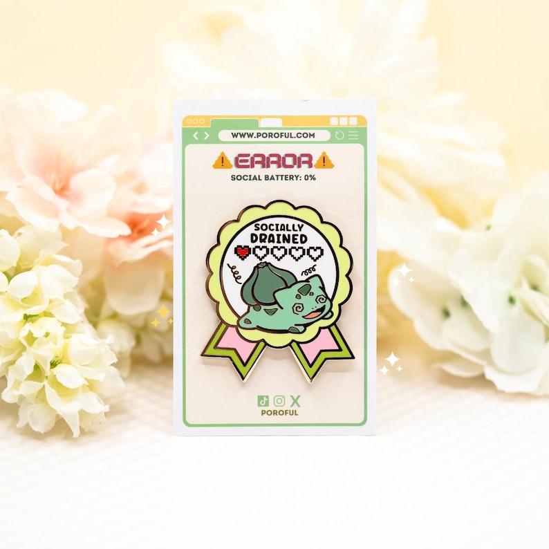 May include: A metal enamel pin shaped like a scalloped award ribbon with pink and light green accents. It features a green creature illustration, the text "SOCIALLY DRAINED," and a pixelated heart icon. The pin is mounted on a white display card.