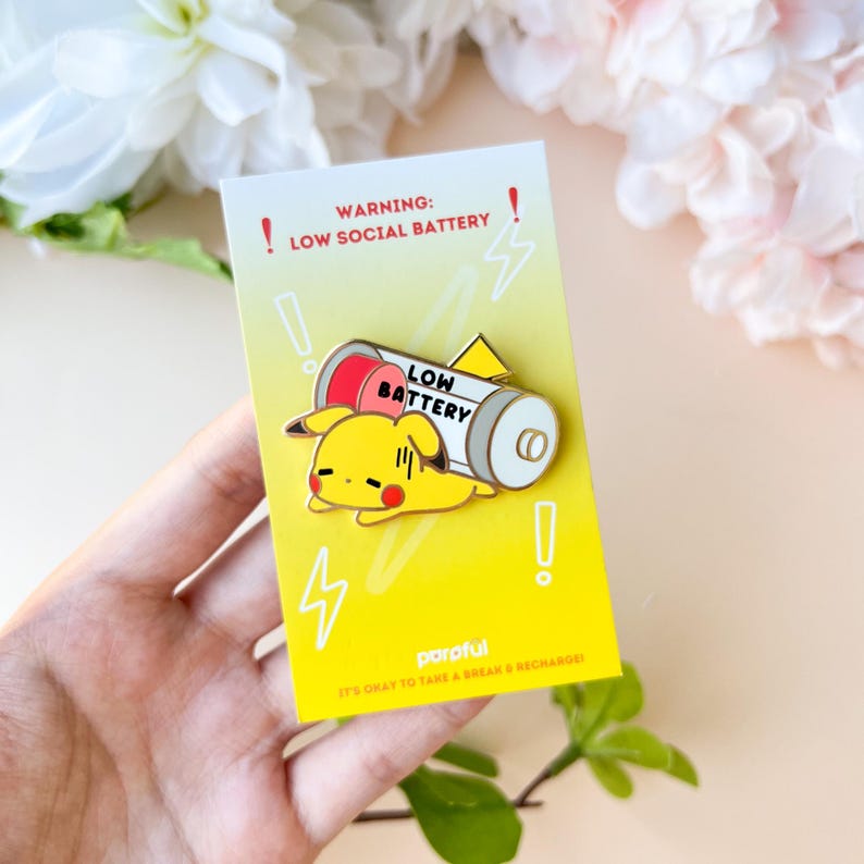 May include: A yellow and white enamel pin featuring a cartoon Pikachu character lying next to a battery with the text "LOW BATTERY". The pin is on a yellow card with the text "WARNING: LOW SOCIAL BATTERY".