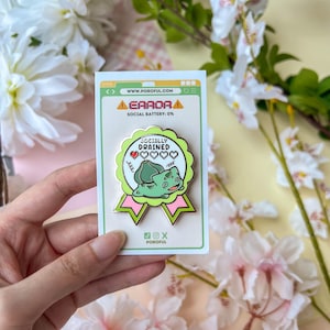 May include: A circular enamel pin with a light green border and pink ribbon tails, featuring a green creature illustration, the text "SOCIALLY DRAINED", and a single red pixelated heart icon. The pin is mounted on a white card with "ERROR" text.