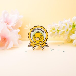 May include: A yellow and white enamel pin shaped like a ribbon with the word "OVERTHINKER" in green text. The pin features a cartoon duck with its hands on its head. The pin is set against a yellow background with flowers.