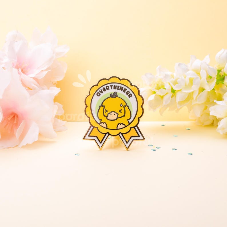 May include: A yellow and white enamel pin shaped like a ribbon with the word "OVERTHINKER" in green text. The pin features a cartoon duck with its hands on its head. The pin is set against a yellow background with flowers.