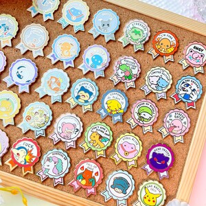 May include: A collection of colorful enamel pins with various designs and phrases. The pins feature cartoon characters and text like "Stressed!", "I'm Fine", and "Dead Inside". Each pin has a ribbon design with a scalloped edge, and is displayed on a corkboard.