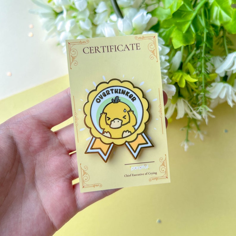 May include: A yellow enamel pin featuring a cartoon duck with the word "OVERTHINKER" in a circular design. The pin is on a yellow certificate card with the text "CERTIFICATE" at the top. The pin has gold-colored accents and a ribbon design.