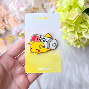 May include: A yellow enamel pin featuring a cartoon Pikachu character lying next to a battery with the words "LOW BATTERY". The pin is on a yellow card with the text "Enamel Pin" and the brand name "parof&uuml;l".