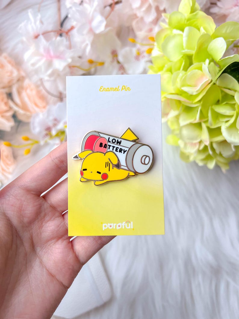 May include: A yellow enamel pin featuring a cartoon Pikachu character lying next to a battery with the words "LOW BATTERY". The pin is on a yellow card with the text "Enamel Pin" and the brand name "parof&uuml;l".