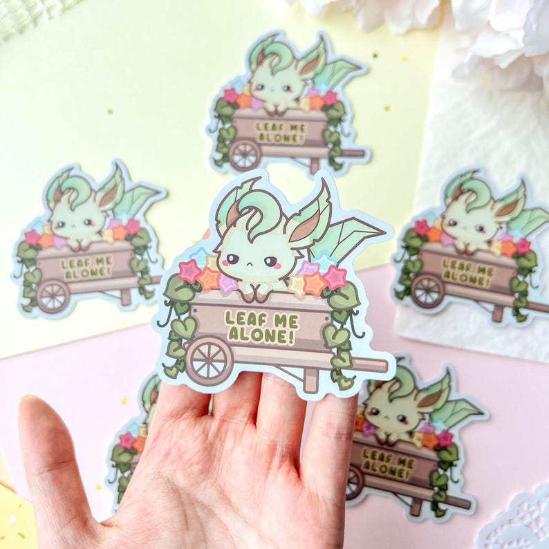 May include: A collection of stickers featuring a cartoon creature with green and cream fur, large ears, and a leafy design. The creature is in a wooden cart with the text "LEAF ME ALONE!" surrounded by flowers and stars.