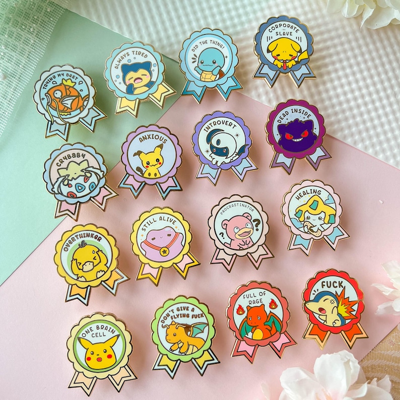 May include: A set of 15 enamel pins featuring various Pokemon characters with humorous phrases. The pins are gold-colored and have a ribbon design. The phrases include "Always Tired", "Did the Thing", "Corporate Slave", "Crybaby", "Anxious", "Introvert", "Dead Inside", "Overthinker", "Still Alive", "Procrastinator", "Healing", "One Brain Cell", "Don't Give a Flying Fuck", "Full of Rage", and "Fuck".