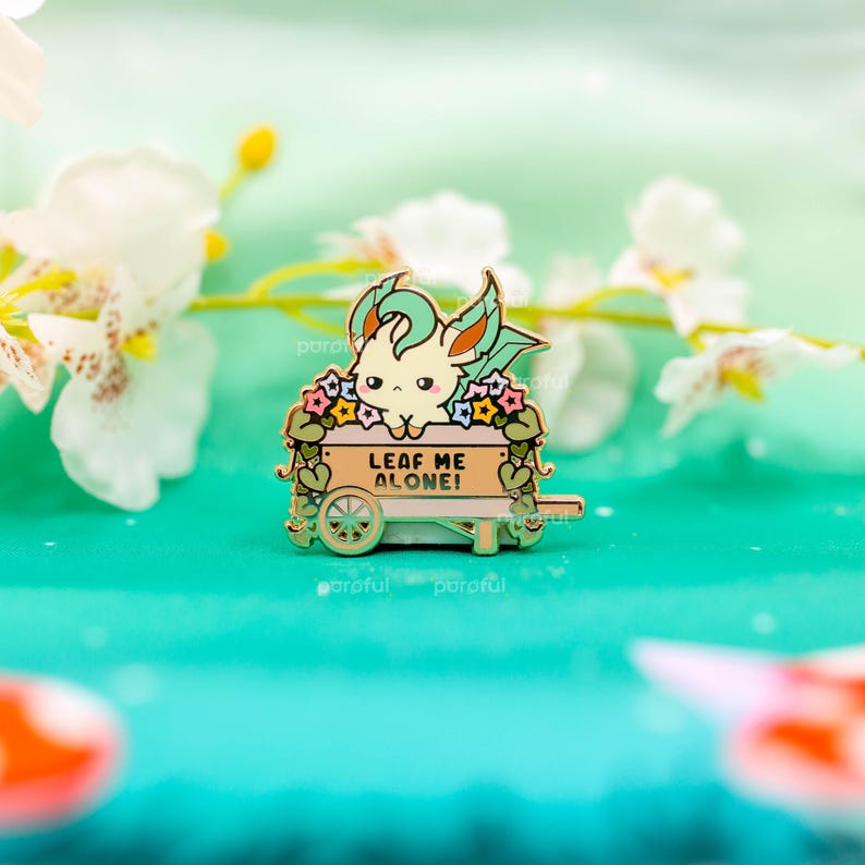May include: An enamel pin depicting a cartoon character in a wooden cart adorned with flowers. The pin reads "LEAF ME ALONE!" and is set against a teal backdrop, with white flowers in the background.