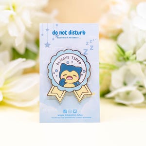 May include: A gold enamel pin with a blue and yellow ribbon. The pin features a cartoon character with a blue body and yellow eyes, sleeping with a mouth open. The text on the pin reads "Always Tired" and "Do Not Disturb, Sleeping in Progress... ZZZ".