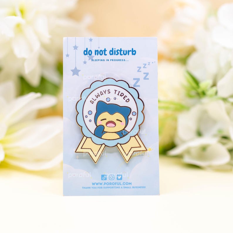 May include: A gold enamel pin with a blue and yellow ribbon. The pin features a cartoon character with a blue body and yellow eyes, sleeping with a mouth open. The text on the pin reads "Always Tired" and "Do Not Disturb, Sleeping in Progress... ZZZ".