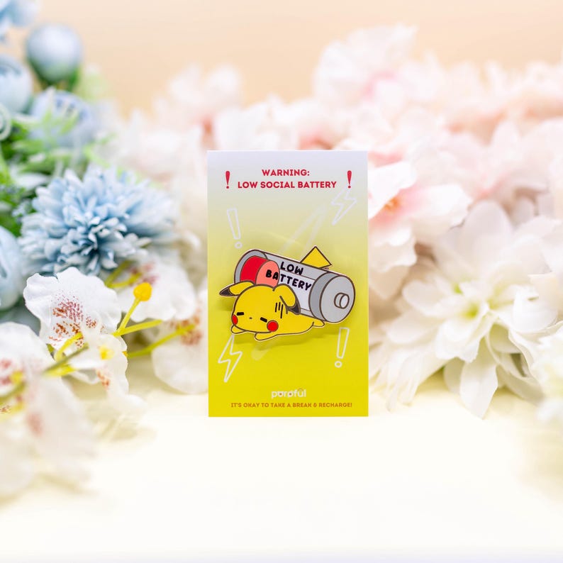 May include: A yellow and red pin featuring a cartoon character with the text "WARNING: LOW SOCIAL BATTERY!" and "LOW BATTERY". The pin is on a yellow background with lightning bolt graphics and the text "IT'S OKAY TO TAKE A BREAK & RECHARGE!"
