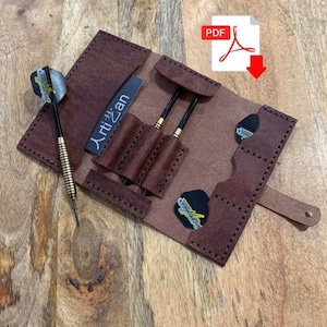 May include: A brown leather dart case with a flap closure and a strap. The case has three slots for darts and two pockets for dart flights. The case has the text "Artisan" printed on the front.