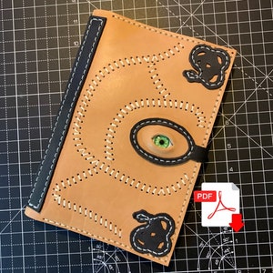 May include: A brown leather journal with a black eye and two black ears sewn on the front. The journal has white stitching around the edges and a black spine. The journal is on a black and white grid background. The text "PDF" is in red on a white background with a red arrow pointing down.