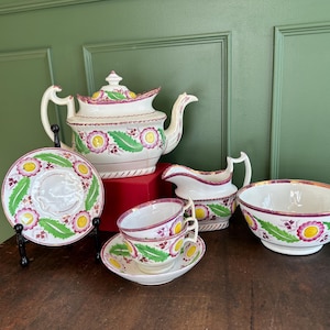 Könnte beinhalten: Ein Vintage-Teeservice mit Teekanne, Sahnekännchen, Zuckerdose, Tassen und Untertassen. Das weiße Porzellanset hat ein Blumendesign mit rosa, gelben und grünen Akzenten. Das Set wird auf einem Holztisch präsentiert.