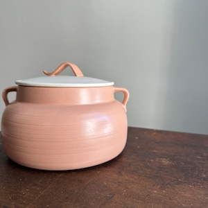 Peut inclure: Un pot en céramique rose avec un couvercle blanc et une poignée incurvée. Le pot a une surface texturée et est posé sur une table en bois.