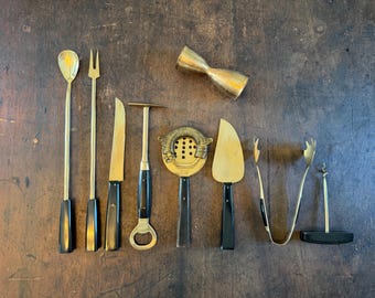 Op de afbeelding: Een set van acht goudkleurige baraccessoires met zwarte handvatten, waaronder een lange lepel, een vork met twee tanden, een mes, een flesopener, een zeef, een schepje, een ijstang en een kurkentrekker, plus een dubbelzijdige jigger.