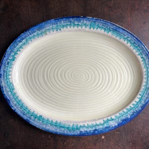 May include: Oval ceramic serving platter with a cream-colored center and a blue and turquoise painted border. The center has a spiral design. The platter is on a dark brown wooden surface. A decorative piece for serving food.