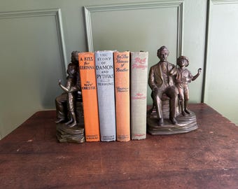 Puede incluir: Cuatro libros de tapa dura antiguos colocados entre dos sujetalibros de metal en tono bronce. Los libros se titulan A Kiss for Corinna, The Story of Damon and Pythias, In the House of Another y Red Pottage. Los sujetalibros muestran figuras humanas.