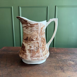 May include: A white ceramic pitcher featuring a brown transferware pattern depicting a rural landscape with a cottage, trees, and a river. The pitcher has a curved handle with a textured geometric design and a wide, flared pouring spout.