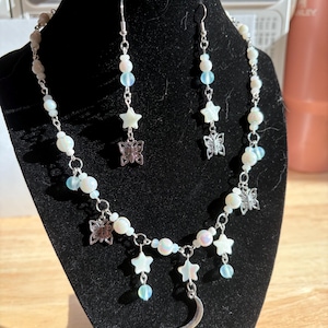 Op de afbeelding: Een sieradenset met een hemels thema, bestaande uit een ketting en oorbellen. De ketting en oorbellen zijn versierd met witte kralen, iriserende blauwe kralen, sterbedels, vlinderbedels en een maanvormige hanger.