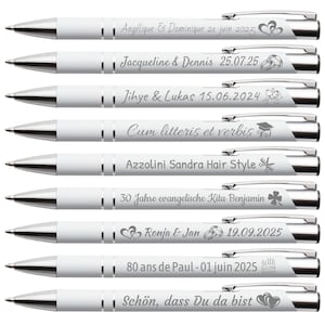 Peut inclure: Plusieurs stylos blancs avec des détails argentés. Chaque stylo est gravé avec un texte personnalisé et des icônes décoratives. Les stylos présentent des noms, des dates et des phrases, notamment "Angélique & Dominique", "Jacqueline & Dennis" et "Schön, dass Du da bist".