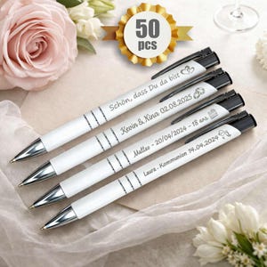 Op de afbeelding: Witte balpennen met zwarte accenten en zilveren punten. Elke pen is gepersonaliseerd met namen en data. Een gouden badge geeft "50 stuks" aan. De pennen liggen op een zacht, lichtgekleurd oppervlak, met bloemen op de achtergrond.