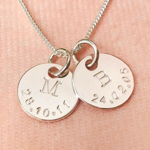 May include: A silver necklace with two round pendants. The pendants are engraved with the letters "M" and "B", and the dates "28.10.11" and "24.02.05".